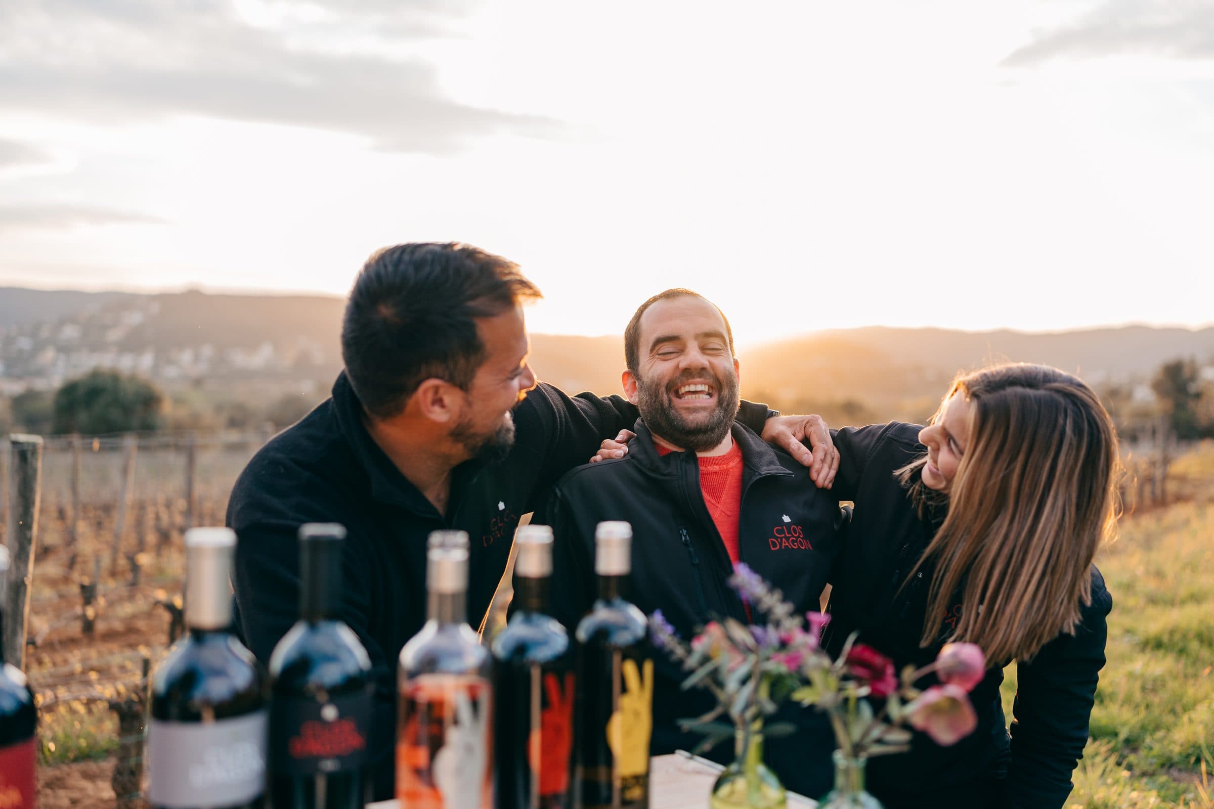 Tres amigos riendo al atardecer con botellas de Clos d'Agon